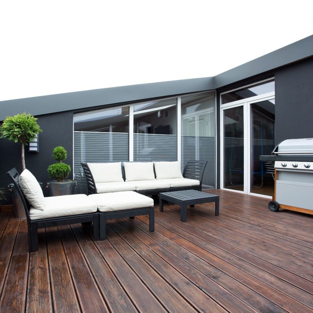Modern outdoor patio with dark exterior walls, wooden decking, sectional seating with white cushions, and a barbecue grill beside large sliding glass doors.