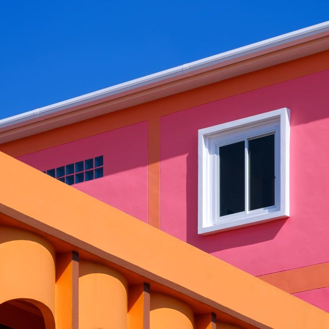 Vibrant minimalist architecture featuring a bright pink facade with orange arched pillars and a white-framed window set against a clear blue sky.