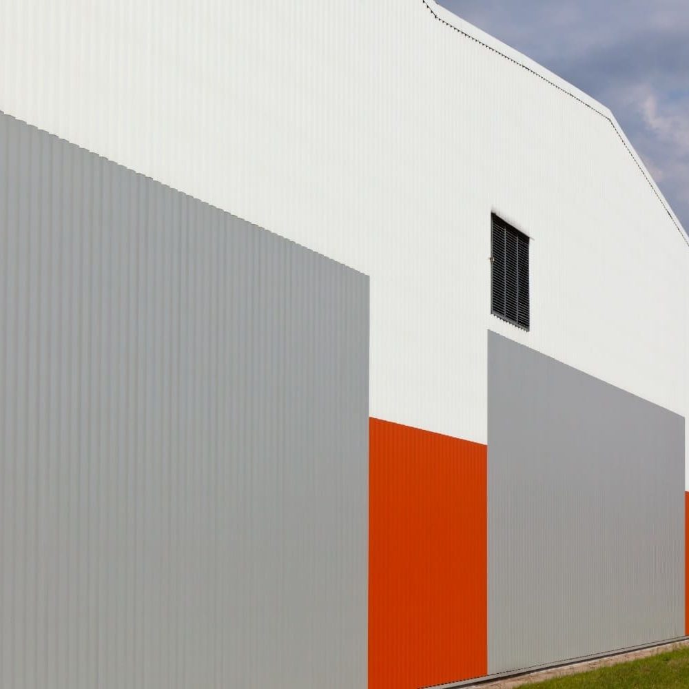 Modern industrial warehouse featuring a geometric facade of corrugated metal panels in white, grey, and bold orange accents under a cloudy sky.