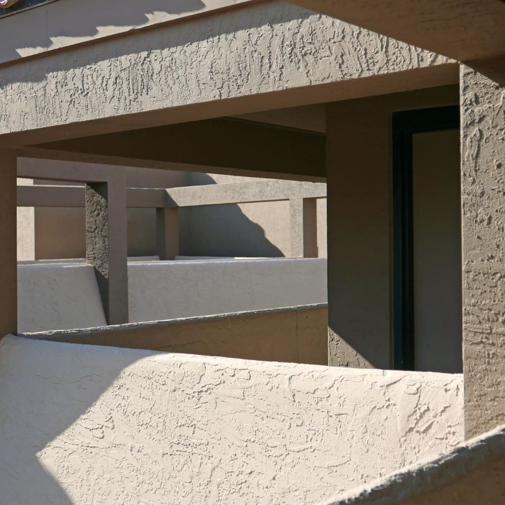 Exterior stucco balcony and walkway with textured beige finish, showing clean architectural lines and sunlit shadows on a residential building.